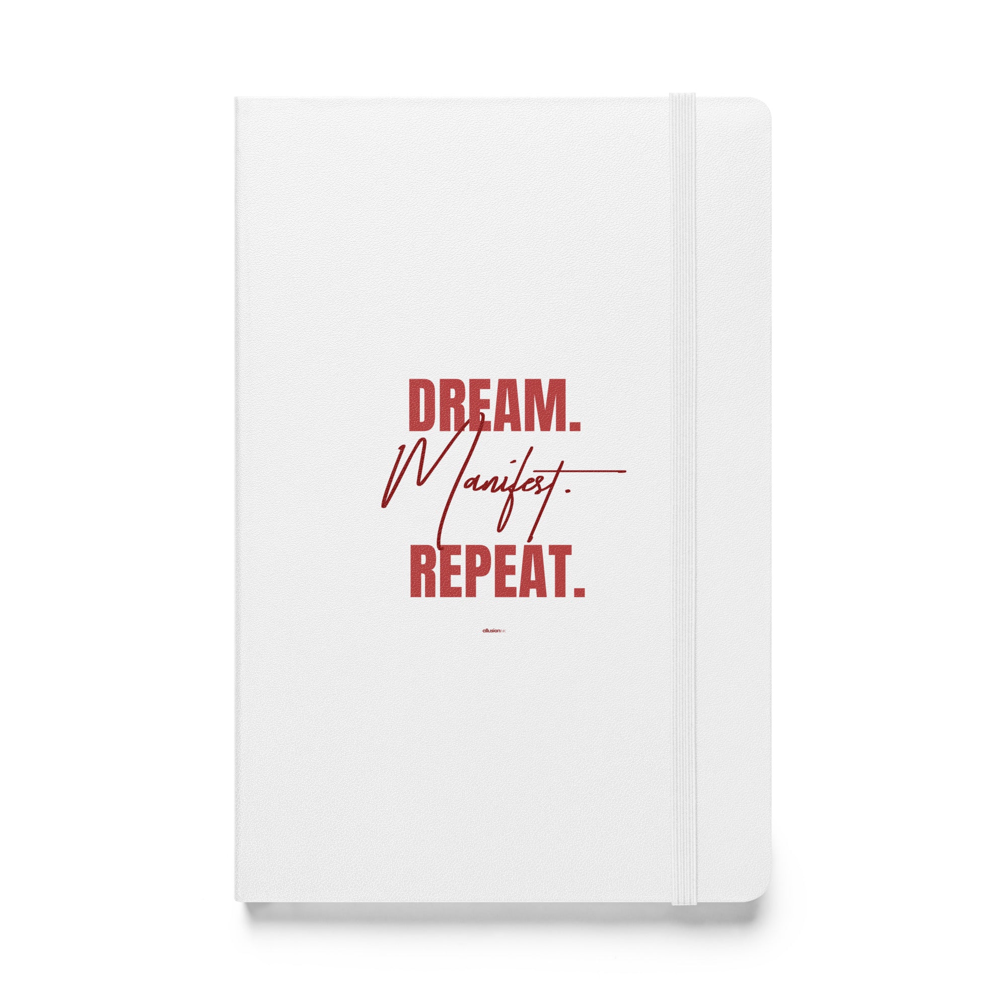 White hardcover notebook featuring the inspiring phrase 'Dream. Manifest. Repeat.' in bold red on the front cover.